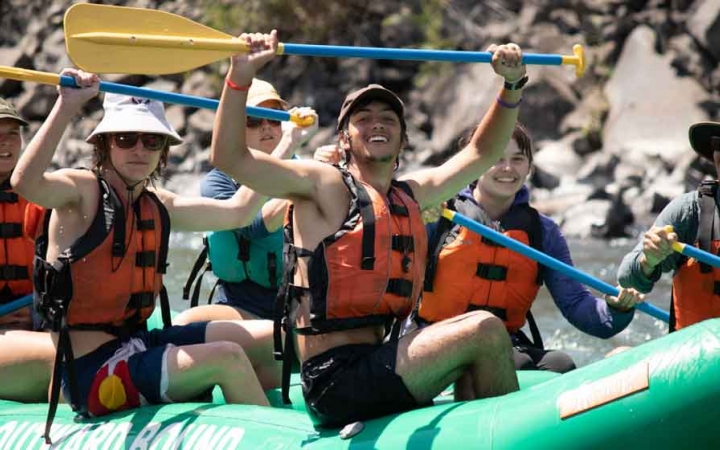 a group of students sitting in a raft smile and raise paddles in the air on an outward bound expedition
